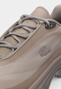 Baskets beiges en tissu mesh, lacets gris, petit logo crocodile sur le côté, et semelle épaisse avec un détail métallique réfléchissant.