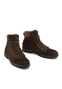 Par de botas de senderismo de ante marrón oscuro con cuellos acolchados en el tobillo, ojales de metal y cordones marrones sobre fondo blanco.