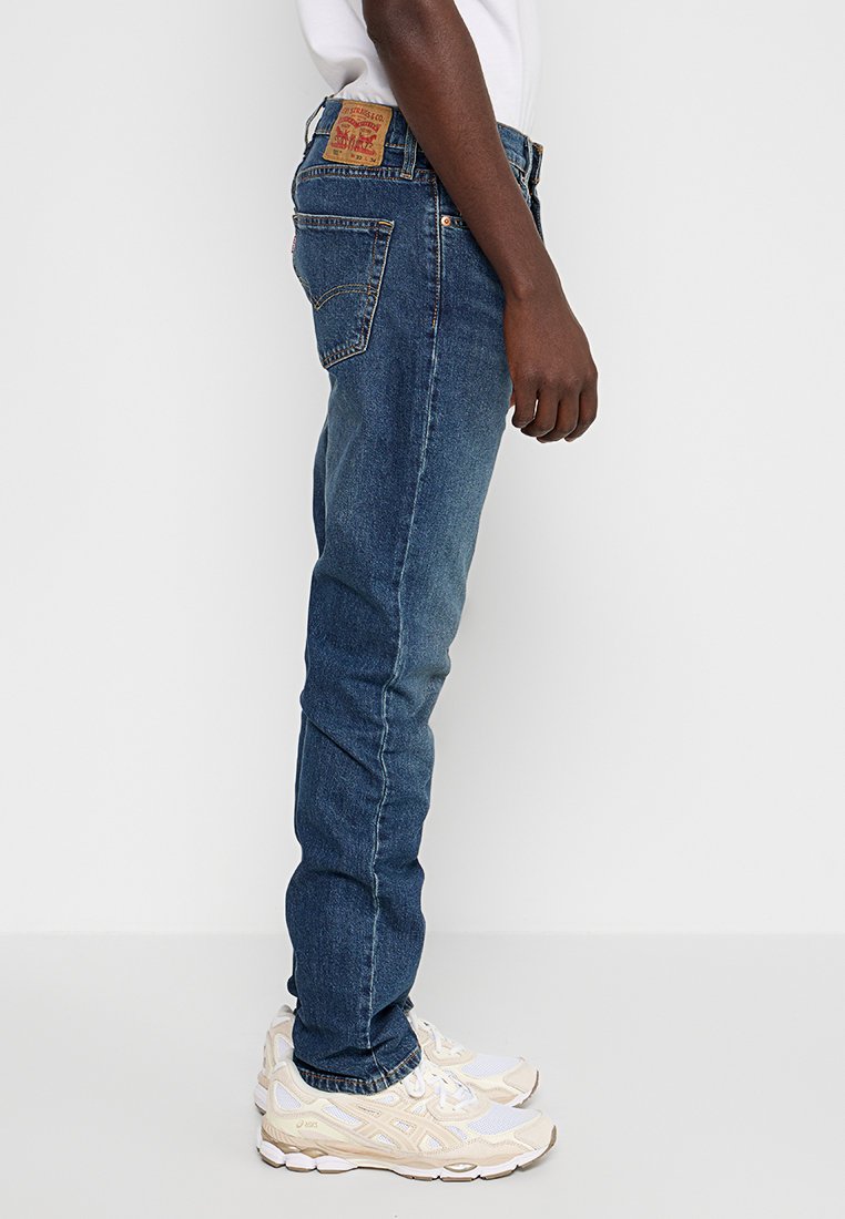 Jean en denim bleu à coupe slim, présentant un design classique à cinq poches et une étiquette en cuir sur la ceinture arrière. Porté avec des baskets de couleur claire.