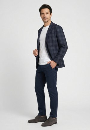 Mann steht und trägt einen marineblau karierten Blazer, weißes T-Shirt, marineblaue Hose und graue Wildleder-Halbschuhe vor einem schlichten hellen Hintergrund.