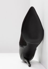 Chaussure noire à talon haut et bout pointu reposant sur la semelle contre un coin blanc entre un mur et le sol, montrant la semelle texturée et le talon fin.