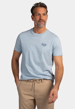 Man met kort haar en baard, gekleed in een lichtblauw NZA New Zealand T-shirt en beige broek met koord, staat met ontspannen handen.