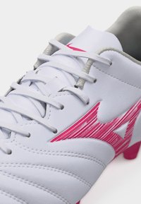 Λευκό ποδοσφαιρικό παπούτσι με ροζ ρίγες στο λογότυπο στο πλάι και λευκά κορδόνια, με επένδυση στη γλώσσα και λεπτομέρειες ραφής εμφανείς.