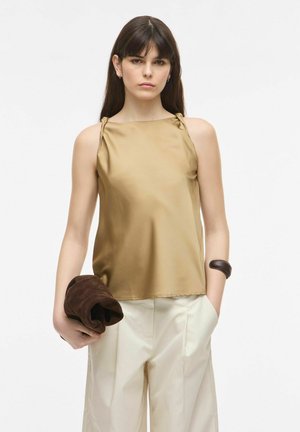 Naine pikkade tumedate juustega kannab varrukateta kuldse topi ja kreemjaid laia säärega pükse, hoides vasakus käes pruunist voltis jopet.