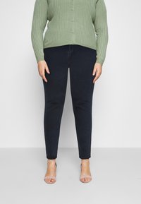 Cardigan vert côtelé à boutons porté sur un jean skinny noir, associé à des talons strappy rose clair. Accent sur la texture et la silhouette ajustée.
