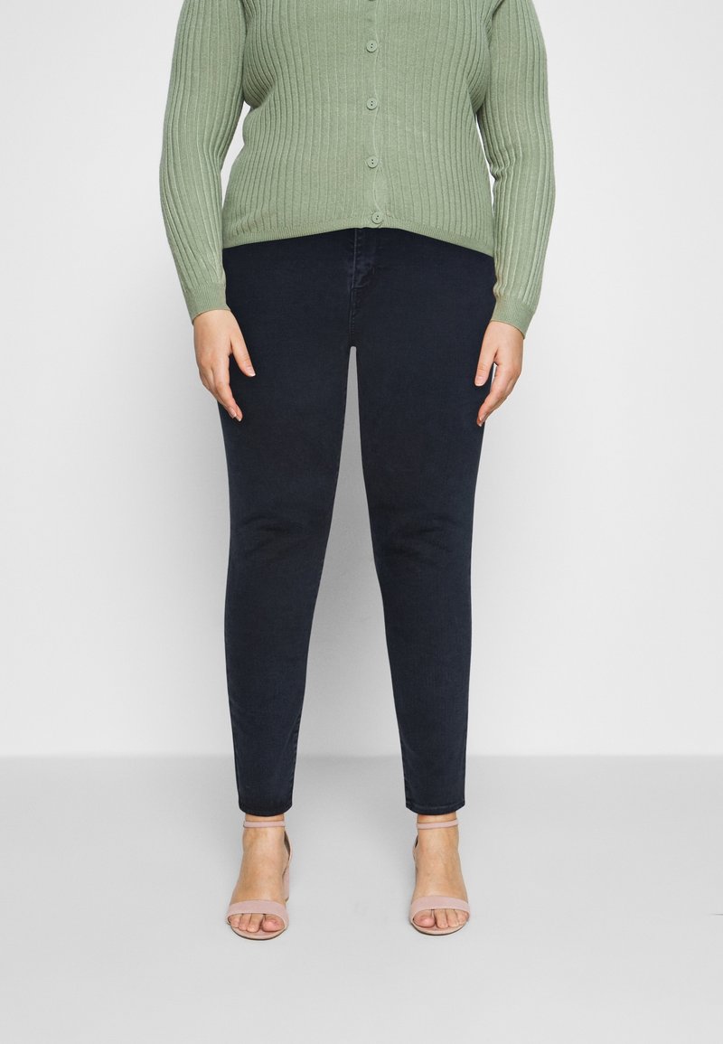 Cardigan vert côtelé à boutons porté sur un jean skinny noir, associé à des talons strappy rose clair. Accent sur la texture et la silhouette ajustée.