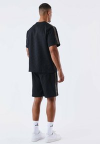 Black textured short-sleeve shirt with patterned side accents, paired with matching shorts. Worn with white sneakers and socks.