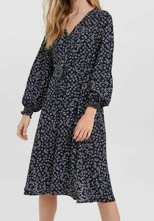 Kvinde iført en navyblå midi-kjole med hvidt blomsterprint, lange puffede ærmer og en V-udskæring.