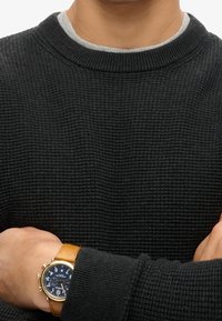 Suéter negro texturizado con cuello redondo gris, combinado con un reloj que presenta una esfera azul y una correa de cuero marrón claro.