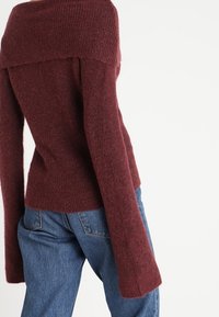 Burgundský svetřík s odhalenými rameny s texturovaným pleteným vzorem a rozšířenými rukávy, kombinovaný s modrými džínami.