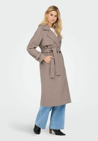 Lang beige trenchcoat lavet af blødt stof, med bælte i taljen, brede revers og to frontlommer, parret med flagrende blå jeans.