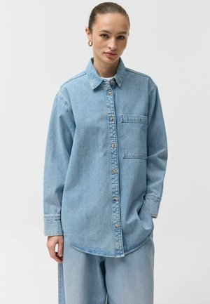 Młoda kobieta z włosami związanymi do tyłu, ubrana w jasnoniebieską jeansową koszulę i dopasowane, luźne spodnie, na tle jednolitej białej ściany.