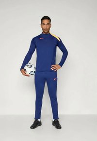 Sininen urheiluasu korkealla kauluksella, keltainen yksityiskohta olkapäällä ja oranssi Nike-logo. Pitelee kuviollista jalkapalloa.