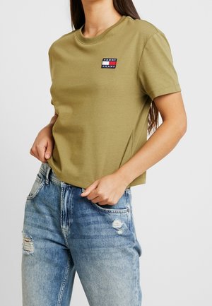 Osoba ubrana w oliwkowozieloną koszulkę Tommy Jeans i jasnoniebieskie, postrzępione dżinsy z rękami blisko pasa.