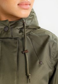 Veste à capuche vert olive avec une finition texturée, dotée de cordons de serrage et de détails en matériel bronze. Gros plan sur le col et les détails de couture.