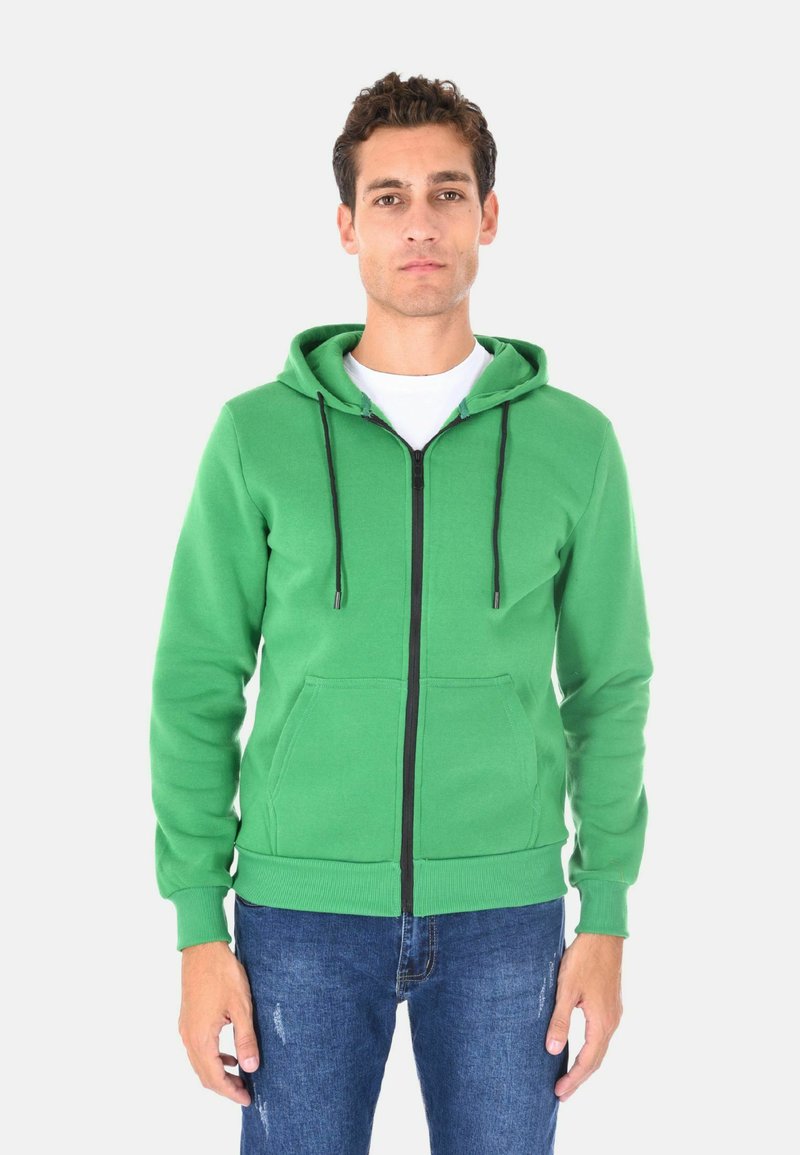 Felpa verde con zip nera, cordini regolabili, tasche frontali e polsini a costine. Abbinata a jeans blu strappati.