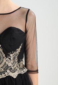 Svart klänning med en genomskinlig illusion halsringning och trekvartsärmar. Guldbroderade detaljer på bodice lägger till ett dekorativt inslag.