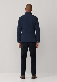 Mand iført en marineblå jakke, sorte bukser og brune sko, som står med ryggen til kameraet mod en ensfarvet lys grå baggrund.