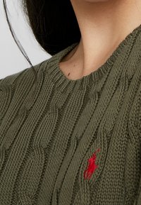 Olivgrüner Strickpullover mit Zopfmuster, Rundhalsausschnitt und einem kleinen roten, gestickten Logo auf der Brust. Die Textur ist dick und strukturiert.