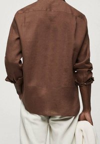 Chemise en lin marron à manches longues, poignets retroussés et coupe décontractée. Le dos présente un ourlet droit et des détails de coutures discrètes.