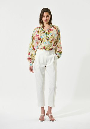 Kleurige blouse met bloemenpatroon en pofmouwen, gecombineerd met witte, hoge geplooide broeken en sandalen met bandjes.