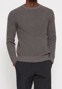 Grauer gestrickter Pullover mit einem gerippten Muster, Rundhalsausschnitt und Raglanärmeln. Mit einem gerippten Saum und Bündchen versehen. Getragen mit schwarzen Hosen.