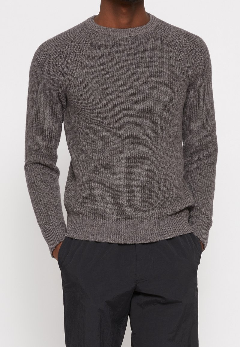 Grauer gestrickter Pullover mit einem gerippten Muster, Rundhalsausschnitt und Raglanärmeln. Mit einem gerippten Saum und Bündchen versehen. Getragen mit schwarzen Hosen.