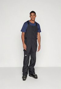 Svarte isolerte kjeledresser med justerbare stropper, knappelukking foran og logodetalj. Brukes over en blå kortermet skjorte.