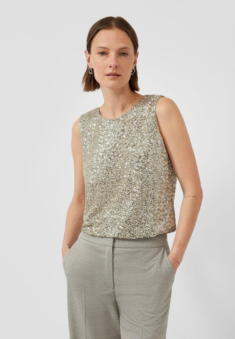 Kobieta z krótkimi brązowymi włosami, ubrana w bezrękawnik z srebrnymi cekinami i jasnoszare spodnie, stojąca z rękami w kieszeniach na jednolitym tle.