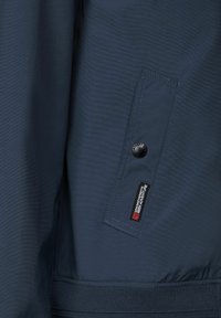 Chaqueta azul marino con un tejido texturizado, bolsillo con cremallera y un botón circular, y puños acanalados. Etiqueta de la marca visible en el bolsillo.