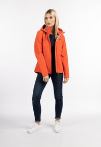 Giacca softshell arancione con cappuccio, chiusura con zip e due tasche frontali, indossata su jeans scuri e sneakers bianche. Texture liscia, design casual.