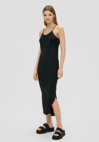 Schwarzes Slipkleid mit dünnen Trägern; schlanker, glatter Textur; knielang mit seitlichen Schlitzen. Kombiniert mit klobigen schwarzen Plateau-Sandalen.