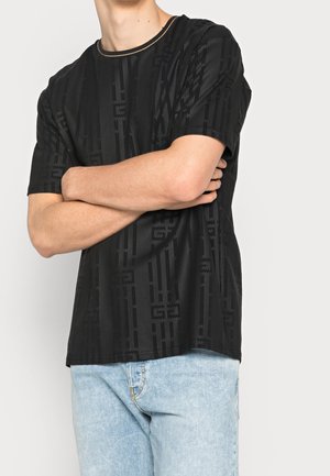 Persona che indossa una camicia a maniche corte nera con motivo e jeans azzurri, con le braccia incrociate, torso visibile su uno sfondo neutro.