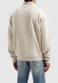 Beige sweatshirt met hoge kraag, geribde manchetten en zoom. Gladde textuur, relaxed fit, van achteren afgebeeld met een blauwe spijkerbroek.