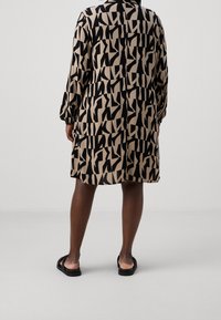 Personne debout, dos à la caméra, portant une robe à manches longues, longueur genou, avec un imprimé abstrait noir et beige marqué et des sandales noires.