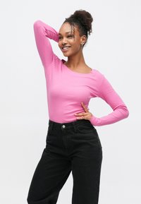 Langärmliges, rosa, geripptes Oberteil mit Rundhalsausschnitt, kombiniert mit hoch taillierten schwarzen Jeans. Das Modell posiert mit einer Hand in der Hüfte und der anderen im Haar.