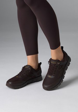 Person trägt schwarze Sportsneaker und schwarze Leggings und steht auf einer hellgrauen Oberfläche.