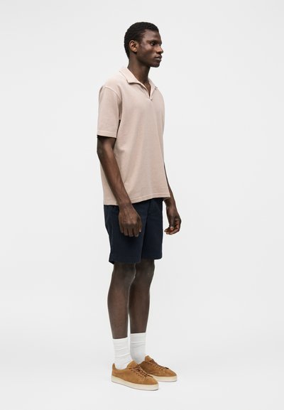 Homme portant un polo beige à manches courtes, un short bleu marine, des chaussettes blanches et des baskets marron, debout de profil sur fond blanc.