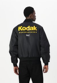 Giubbotto bomber nero con un grande logo giallo "Kodak" sul retro, con polsini e vita a coste neri e una texture liscia e lucida.