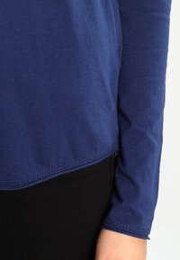 Sininen, pitkähihaisen puuvillapaita, jossa on pyöreä helma ja näkyviä ompelutietoja, yhdistettynä mustiin housuihin.