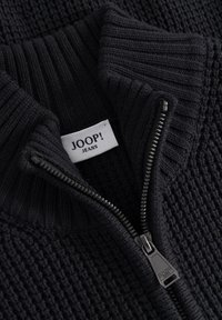 Czarny sweter z dzianiny z częściowo rozpiętym czarnym zamkiem i białą metką "JOOP! JEANS" naszytą wewnątrz kołnierza.