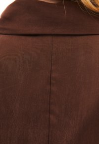 Hnědá texturovaná tkanina s hladkým, mírně lesklým povrchem. Obsahuje středový šev probíhající vertikálně podél zadní části.