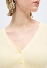 Γυναίκα με κοντά μαλλιά που φοράει χρυσούς κρίκους σκουλαρίκια και ανοιχτό κίτρινο τοπ με V-λαιμόκοψη και κουμπιά, με ορατή περιοχή λαιμού και κλείδας.