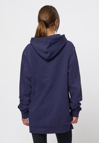 Personne aux cheveux bruns attachés en queue de cheval, portant un sweat à capuche ample bleu foncé et un pantalon noir, de dos devant un fond uni.