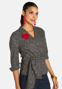 Kurzärmlige marineblaue Bluse mit einem goldenen Wirbelmuster, an der Taille gebunden, mit einem roten Blumenakzent und getragen mit goldenen Armreifen.
