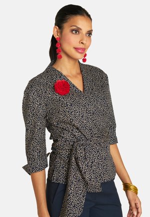 Kurzärmlige marineblaue Bluse mit einem goldenen Wirbelmuster, an der Taille gebunden, mit einem roten Blumenakzent und getragen mit goldenen Armreifen.