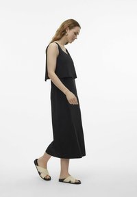 Black sleeveless dress with a flowy silhouette and a cropped top layer. Paired with cream cross-strap sandals featuring black soles.