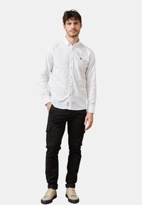 Camisa blanca de botones con un pequeño logo en el pecho, combinada con pantalones cargo negros y zapatillas beige. Modelo de pie frente a un fondo plain.