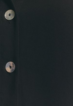 Tessuto nero con una leggera texture, caratterizzato da un grande bottone rotondo e lucido. Il bottone ha un solo foro al centro.