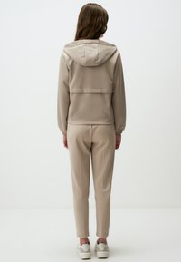 Giacca a vento beige con cappuccio e pantaloni affusolati abbinati in un tessuto morbido e liscio. Presenta polsini elasticizzati e un design minimalista. Sneakers bianche completano il look.